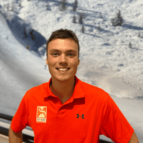 Een enthousiaste instructeur in een rood ski- of snowboardshirt, met een besneeuwd berglandschap op de achtergrond.