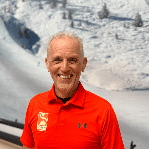 Man in oranje ski jas met Ski-Mere logo, glimlachend met besneeuwde bergen op de achtergrond.
