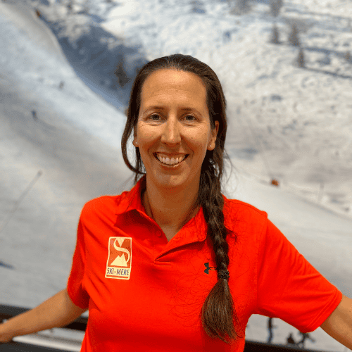 Skihandelaarvrouw in rode ski-instructeurtenue met skis in indoor skibaan in Almere, lachend en klaar voor lessen.