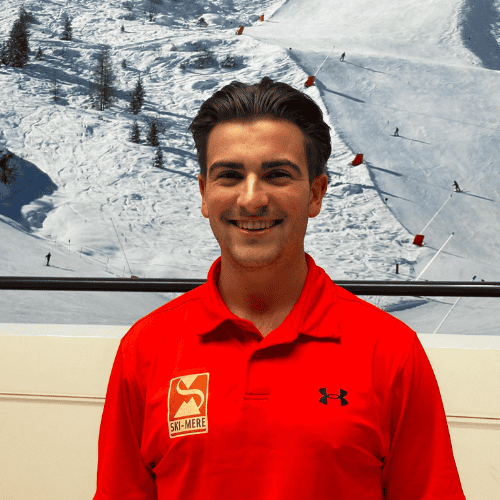 Ski-instructeur op indoor rolpiste in Almere, lachend en in rood ski-jasje, met sneeuwrijke bergachtergrond.