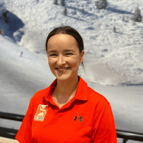 Vrouw in oranje ski- of snowboardoutfit met sneeuwrijke bergen op de achtergrond.