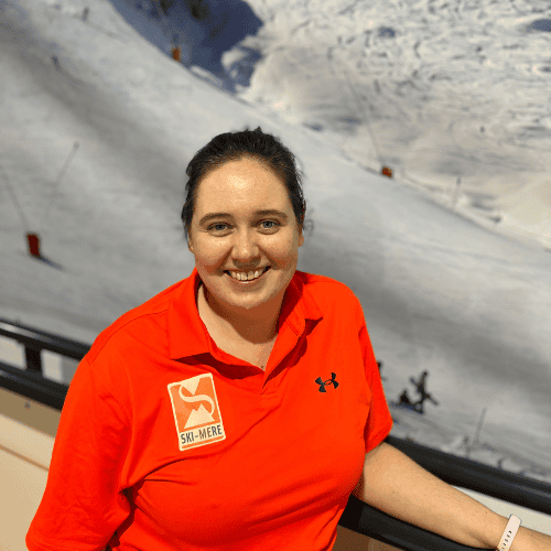 Sneeuwrijke skibaan met instructeur in rode jas, indoor ski- en snowboardlessen in Almere, glimlachende instructeur met skiplezier op de achtergrond binnen rolpiste.