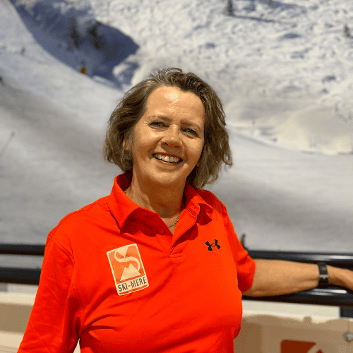 Ski-instructeur met grijze haar en rode sportkleding, lachend voor een sneeuwachtergrond met bergen en een blauwe lucht.