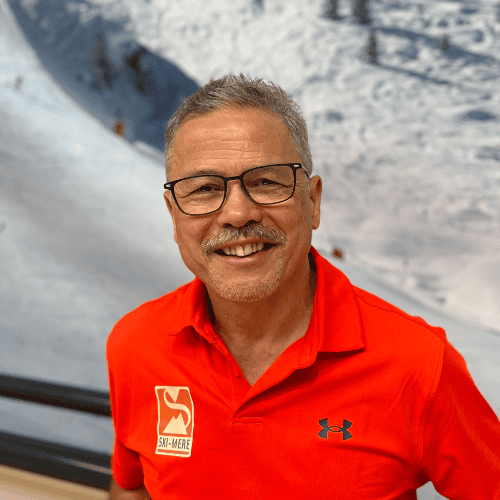 Man in rode ski-ondernemings polo met ski-mere logo, voor een indoor skipiste in Almere, glimlachend, in de sneeuw, winteratmosfeer.