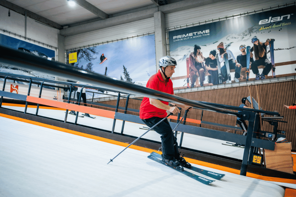 Skileraar op de indoor rolpiste in Almere, met posters van ski- en snowboardmerken op de muur.