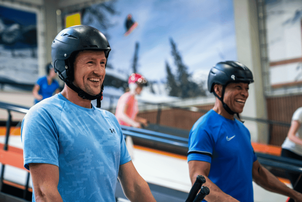 Kleine groep mensen met helmen op, lachend en plezier hebbend op een indoor ski- en snowboardrolpiste in Almere, met een winterlandschap op de achtergrond.