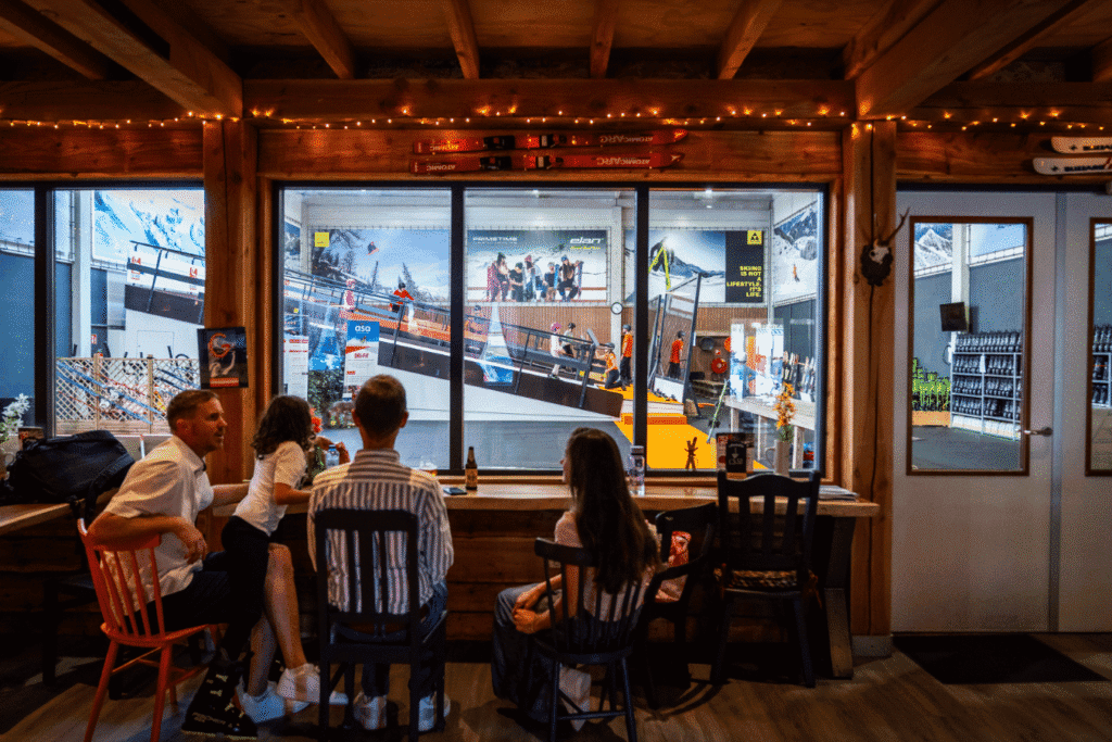 Sfeervol interieur van een skihut of café met uitzicht op de indoor rolpiste, waar mensen genieten van hun drankje terwijl ze naar de skilessen en -activiteiten kijken.