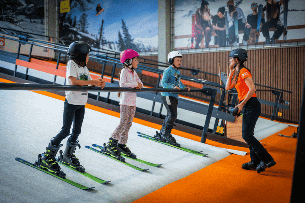 Ski- en snowboardlessen op een indoor rolpiste in Almere, met instructrice die kinderen begeleidt tijdens een les. Kinderen dragen helmen en skis, klaar voor een leuke en leerzame ervaring.